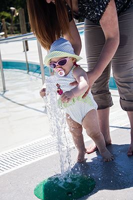 Infant with sunhat and sunglasses playing in fountain while being held by an adult